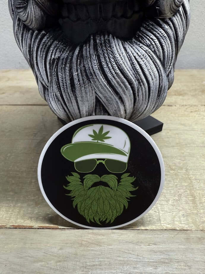 Weed Beard Man 3-Inch Sticker featuring a bearded character with cannabis design and sunglasses on a wooden table.