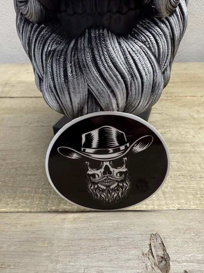 Cowboy Beard Skull sticker showcasing a skull with a beard and cowboy hat, placed on a rustic wooden surface.