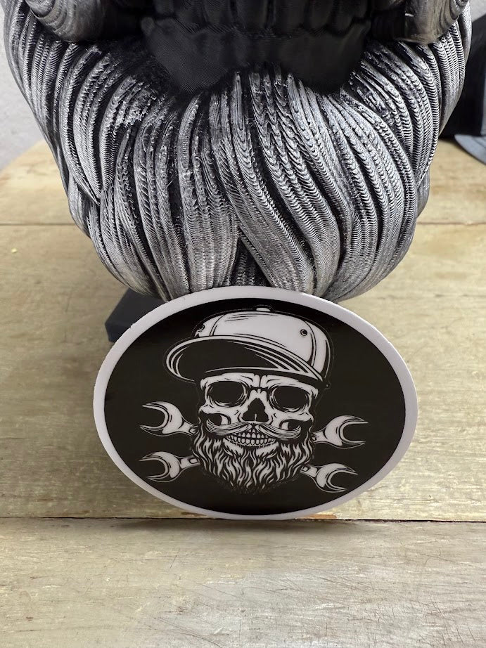 Bearded skull mechanic sticker on a wooden surface, featuring a rugged design with a cap and wrenches.